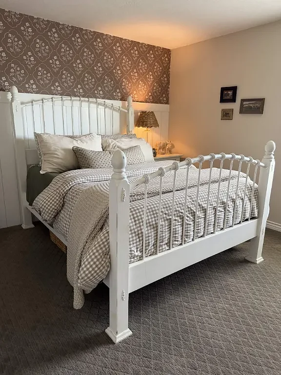 The finished bedroom: white vertical boards to a cap rail with the floral paper above them, a made-up bed with a white iron frame against the wall, and a lit lamp on the nightstand