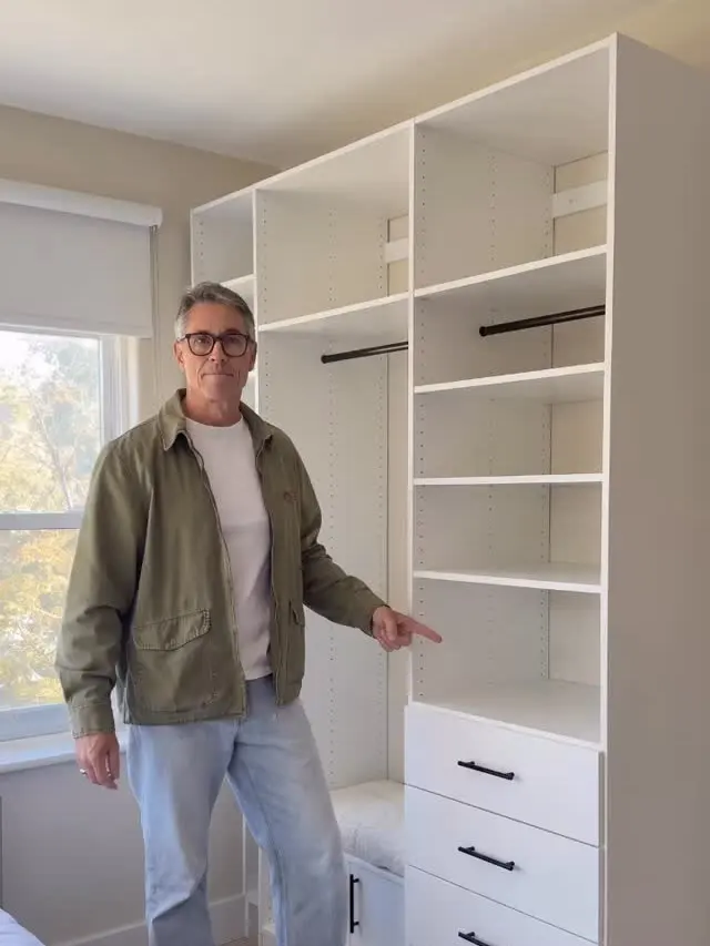 Russ standing in the finished walk-in: white shelving to the ceiling, 2 hanging rails, a bank of drawers with black pulls and a bench seat beside the window