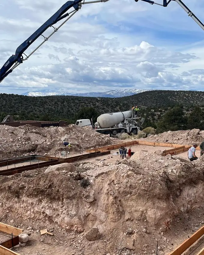 A concrete pump boom arcing over formed and steeled footings, the truck and mixer parked against the snow-topped mountains