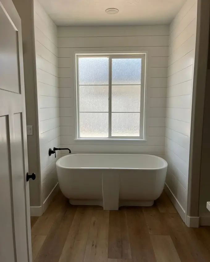 A finished bathroom with a freestanding soaking tub under an obscured-glass window, shiplap walls and wide plank flooring