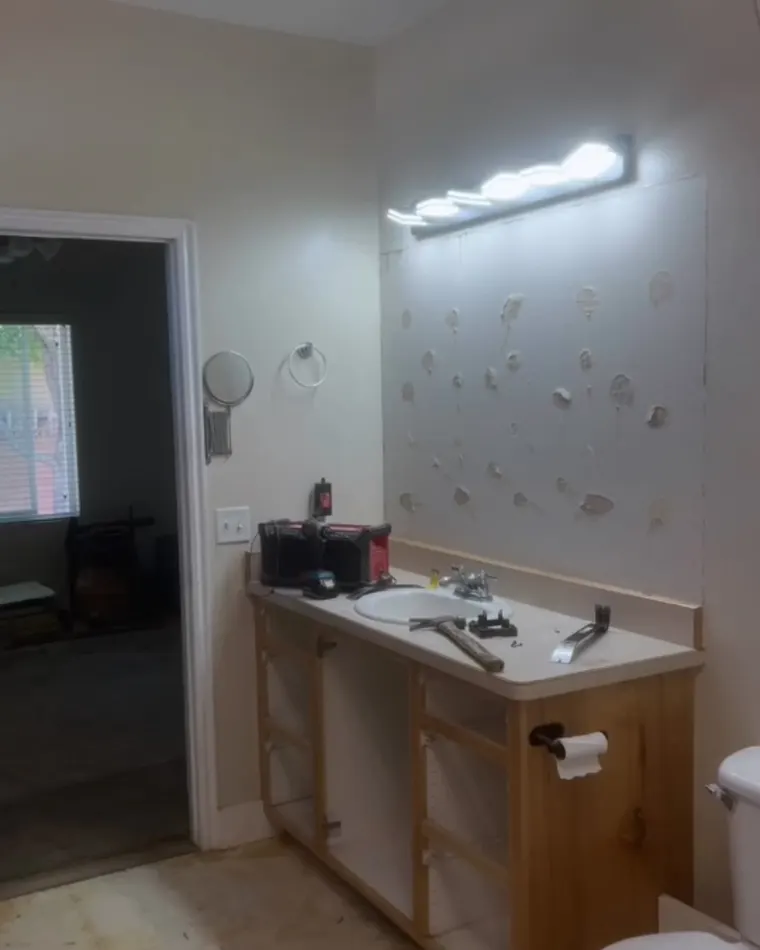The same bathroom during tear-out: the mirror off the wall leaving its adhesive behind, cabinet doors and drawers pulled and tools laid out on the counter